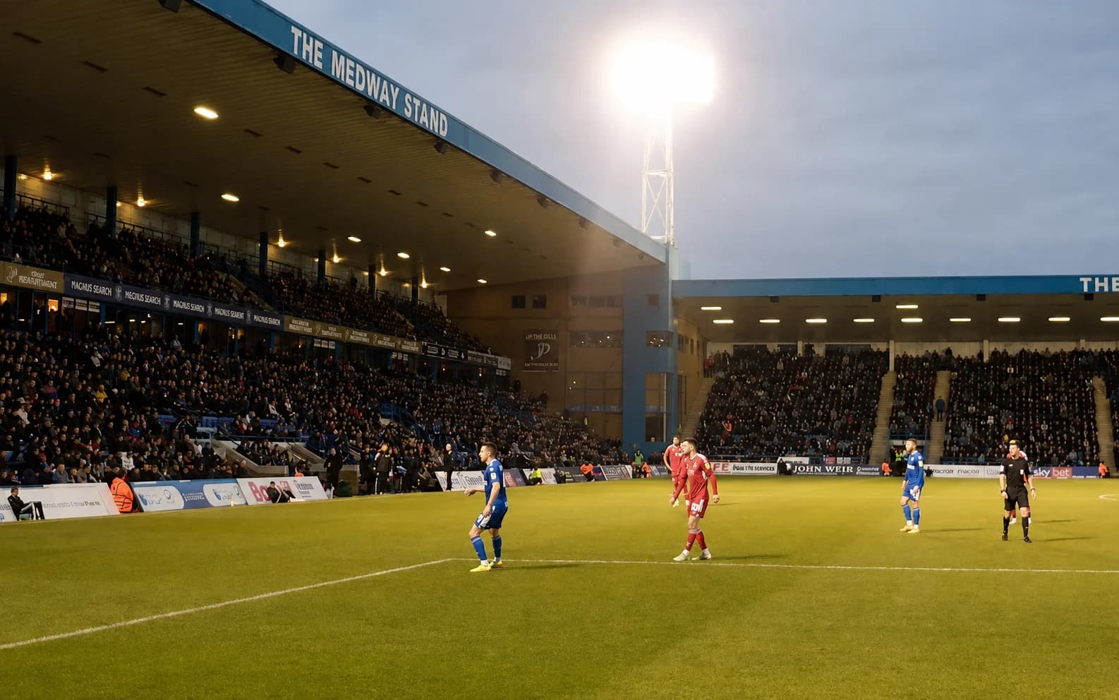 Football match under floodlights at Priestfield Stadium, players in blue and red kits on the pitch, a stand full of spectators in the background.